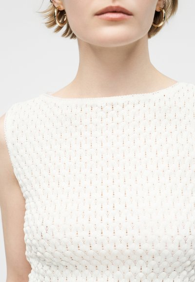 Κοντινό πλάνο ενός ατόμου που φοράει λευκό υφασμάτινο αμάνικο τοπ και μεγάλα χρυσά σκουλαρίκια κρίκους, εμφανίζοντας το κάτω μέρος του προσώπου και τον λαιμό.