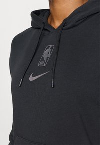 Nærbilde av en person som bærer en svart hettegenser med reflekterende NBA- og Nike-logoer på brystet og svarte snorer.