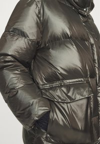 Błyszcząca ciemna kurtka puchowa z dużą klapką kieszonki i widoczną dłonią wsuniętą do kieszeni, pokazująca pikowane przeszycia i błyszczącą teksturę tkaniny.