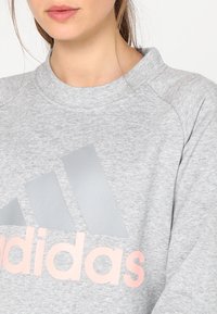 Femme portant un sweat-shirt Adidas gris clair avec un grand logo argenté et des lettres roses pâles sur la poitrine, cadré au niveau de la bouche et des épaules.