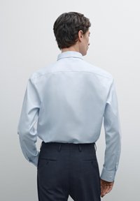 Lichtblauw, overhemd met lange mouwen en een achterpand, met knopenmanchetten, gedragen met donkere broeken. Glad stof zonder patronen of accenten.