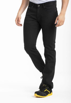 Jean en denim noir avec une coupe slim, un design à cinq poches, et des détails de couture visibles, associés à des chaussures de sport noires et jaunes.