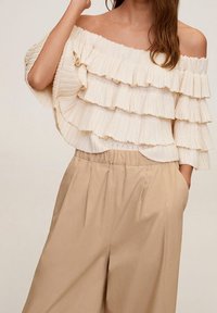 Vrouw in beige wijde broek en een off-shoulder crèmekleurige gebreide top met gelaagde ruches, poseert met één hand in de zak.