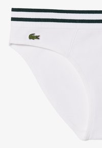 Culotte blanche côtelée avec ceinture rayée verte et blanche et petit logo crocodile vert sur le devant gauche.