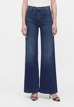 Femme portant un jean bleu foncé taille haute, jambes larges, avec des poches plaquées à l'avant et des talons ouverts, debout devant un fond uni.
