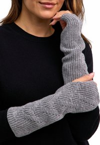Vrouw met lange grijze gebreide vingerloze handschoenen met kabelpatroon over een zwart sweater, die met één hand haar gezicht aanraakt.