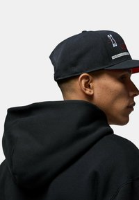 Cappello nero con numeri ricamati 23 e 1989 sulla parte anteriore, visiera interna rossa, abbinato a una felpa nera visibile sullo sfondo.