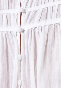 Tessuto bianco leggero con pieghe verticali, caratterizzato da diversi bottoni rivestiti in tessuto lungo il centro e dettagli di cucitura delicati.