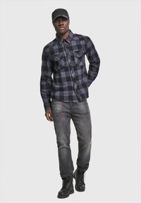 Camisa a cuadros negra y gris con bolsillos en el pecho abotonados, combinada con jeans grises y botas negras con cordones, llevada con una gorra negra.
