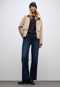 Krämfärgad ulljacka med fickor, mörkgrön tröja, mörkblå vida jeans och svarta stövlar. Modellen bär en mörk mössa.