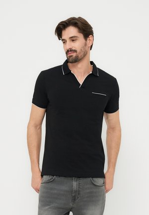 Homme portant un polo noir à manches courtes avec des bordures blanches sur le col, les bords des manches et la poche de poitrine, associé à un jean gris, debout devant un fond blanc.