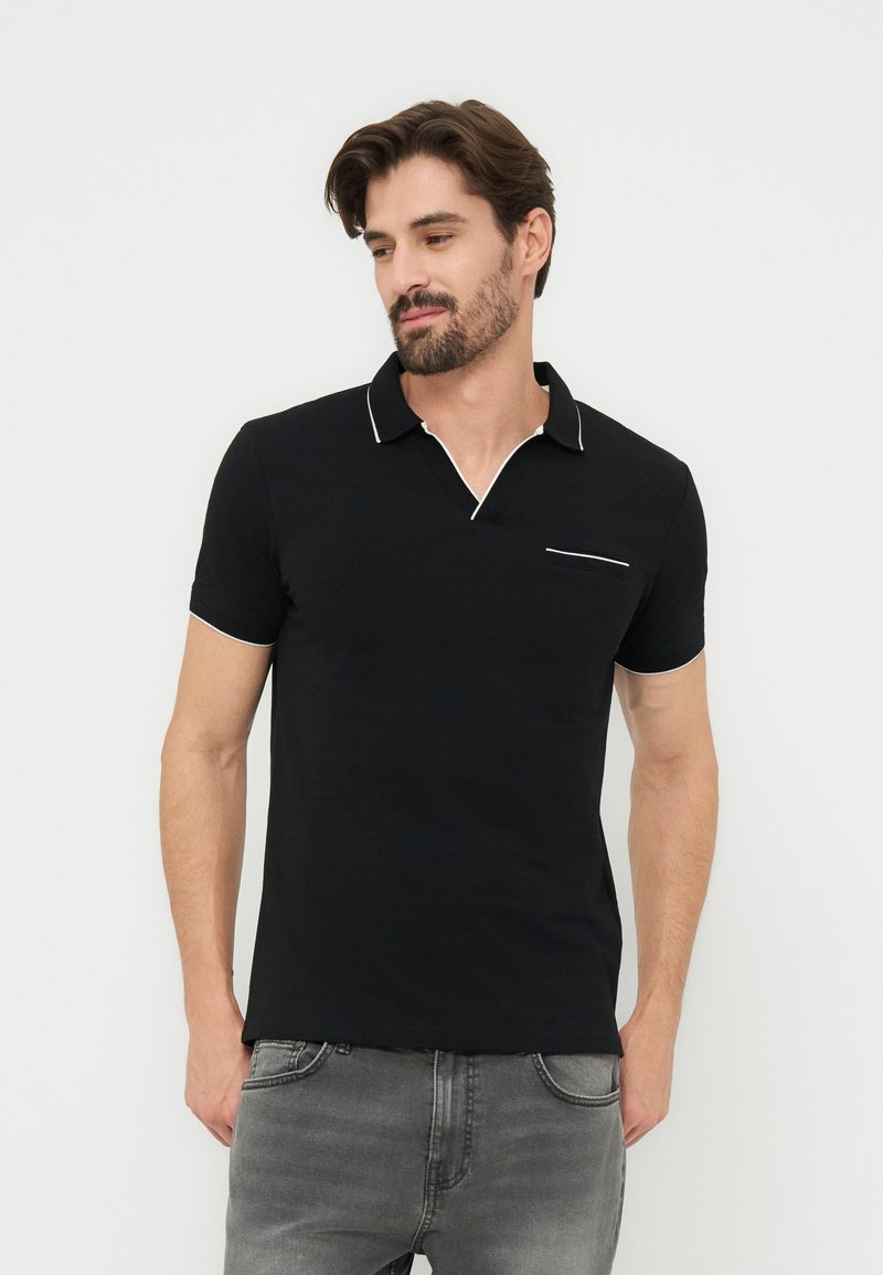 Άνδρας που φοράει μαύρο κοντομάνικο πόλο με λευκή επένδυση στο γιακά, στις άκρες των μανικιών και στην τσέπη στο στήθος, συνδυασμένο με γκρι τζιν, στέκεται μπροστά σε λευκό φόντο.