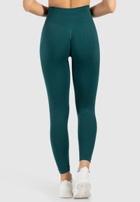 Leggings a vita alta di colore teal realizzati in un tessuto elastico e liscio, con un design senza cuciture e lunghezza alla caviglia, abbinati a sneaker bianche.