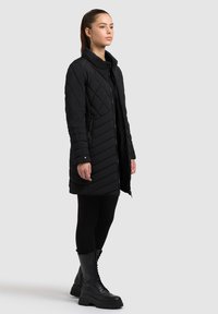 Zwarte gestikte lange jas met een hoge kraag, ritszakken en een gestructureerd patroon. Gecombineerd met zwarte leggings en veterschoenen.