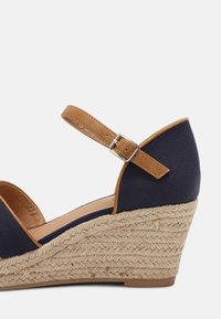 Sandale compensée en toile navy avec une sangle de cheville marron et une boucle argent ajustable. Talon compensé enveloppé de corde de jute pour un détail texturé.