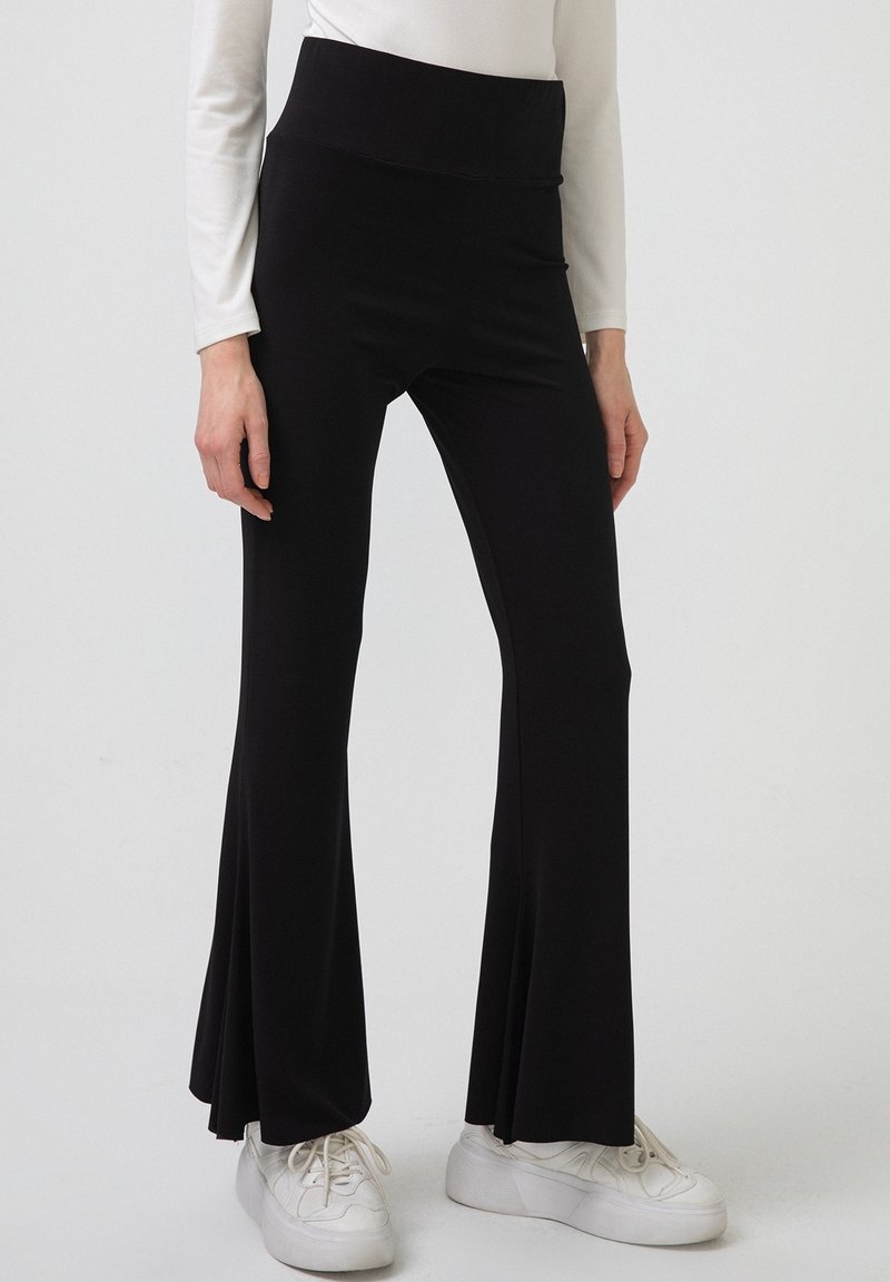 Femme portant un pantalon évasé noir taille haute, un haut blanc à manches longues et des baskets à plateforme blanches, debout devant un fond uni.