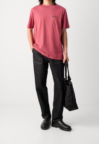 Camiseta de algodón rosa suave con un pequeño logo, combinada con pantalones de mezclilla oscura y botas de cuero negras. Se sostiene un bolso tote negro en la mano.