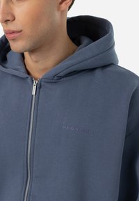 Tmavomodrá mikina na zips vyrobená z mäkkého materiálu, s klokankovou kapsou, veľkou kapucňou a jemným logom na hrudníku. Hladká textúra.