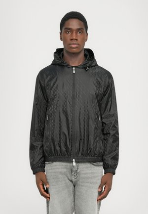 Black hooded jacket with a zip front, diamond-pattern texture, elastic cuffs, and a drawstring hem. Worn over light gray jeans.