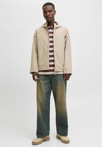 Beige, lett jakke med hette over en stripete, brun og hvit skjorte. Løse, falmede blå jeans og beige støvler fullfører antrekket.