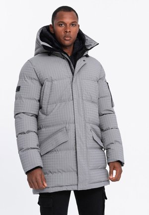 Grijs pufferjack met een gequiltde textuur, grote capuchon, ritssluiting en meerdere zakken, gedragen over een zwart jack.