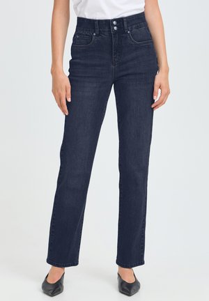 Femme portant un jean droit taille haute bleu foncé avec fermeture à double bouton et chaussures noires pointues.