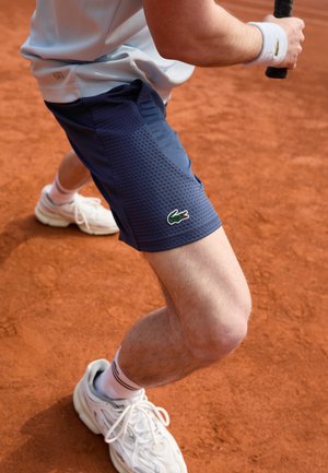 Tennisspieler in marineblauen Shorts und weißen Turnschuhen bereitet sich darauf vor, den Schläger auf einem Sandplatz zu schwingen.