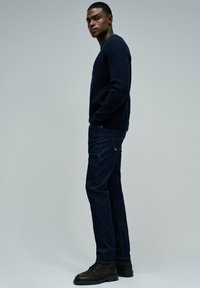 Hombre con un suéter azul oscuro, jeans oscuros y botas negras de pie con las manos en los bolsillos frente a un fondo gris liso.
