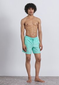 Νεαρός άντρας που στέκεται ξυπόλητος φορώντας μίντ πράσινα μαγιό μπροστά σε απλό ανοιχτό φόντο.