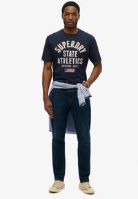 Camiseta de algodón azul marino con texto blanco "Superdry State Athletics" y un pequeño diseño de bandera, combinada con jeans de mezclilla oscura y zapatillas beige.