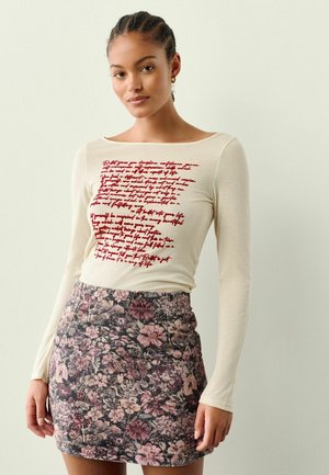 Long-sleeve cream top with red handwritten text, paired with a fitted floral mini skirt in dark tones and pink accents.