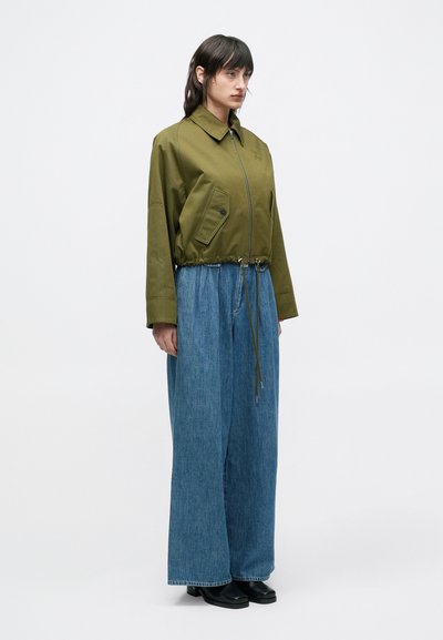 Χακί πράσινο κοντό μπουφάν με φερμουάρ και μπροστινή τσέπη, σε συνδυασμό με μπλε τζιν παντελόνια φαρδιάς γραμμής, με ρυθμιζόμενο κορδόνι στη μέση.