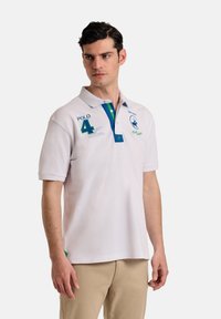 Camisa polo blanca de manga corta, con cuello azul y acentos verdes. Presenta texto bordado y un logo en el pecho. Corte regular.