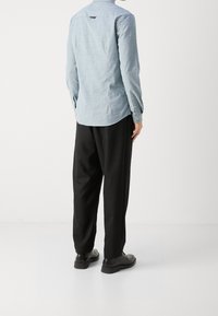 Mies, joka seisoo kulmassa, päällä vaaleansininen pitkähihaispaita, mustat löysät housut ja mustat siistit kengät valkoista taustaa vasten.