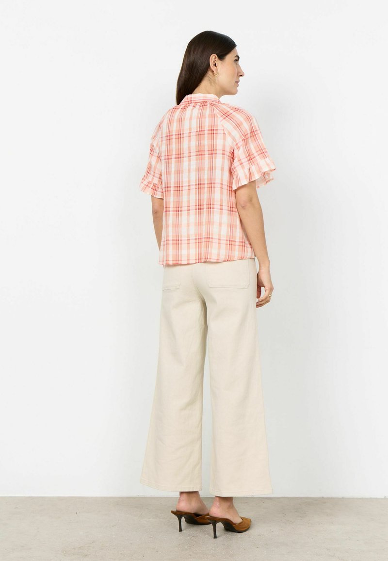 Femme de profil portant un chemisier à manches courtes à carreaux roses et oranges, un pantalon large crème et des mules à talons hauts marron.