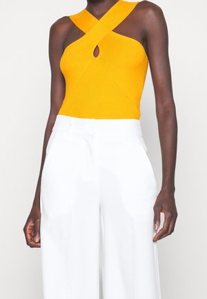 Femme portant un haut tricoté jaune sans manches avec des bretelles croisées et un pantalon blanc taille haute à jambes larges, debout devant un fond uni.