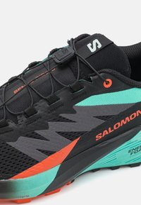 Scarpa da trail running nera con parte superiore in mesh, accenti turchesi e arancioni, dotata di sistema Quicklace, suola Contagrip e elementi di design testurizzati.