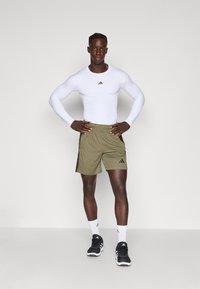 Prigludusi balta ilgomis rankovėmis maikė, alyvuogių žalios šortai su juodomis šoninėmis juostelėmis, juodi sportbačiai ir balti kojinės su juodais akcentais.