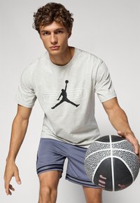 Camiseta gris claro con el logo negro de Jumpman, combinada con pantalones cortos deportivos azules con rayas negras. El modelo sostiene un balón de baloncesto texturizado.
