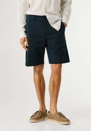 Man in een beige shirt met lange mouwen, marineblauwe knielange korte broek en lichtbruine casual veterschoenen, staand met één hand in de zak.