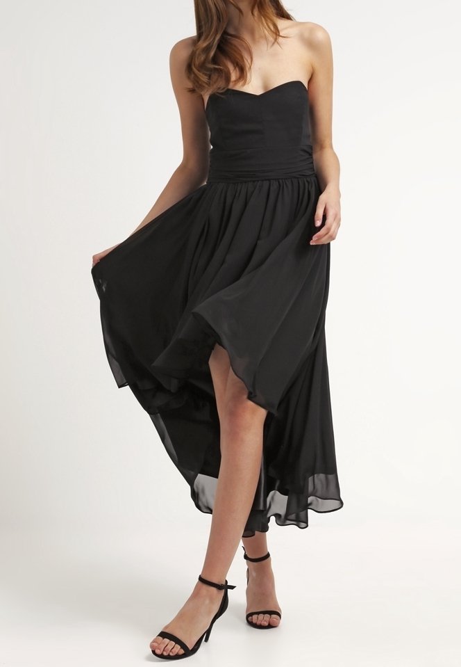 Femme portant une robe noire sans bretelles avec un ourlet haut et bas et des talons à bride cheville noirs, tenant sa jupe pour dévoiler ses jambes devant un fond uni.