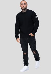 Zwarte geribbelde gebreide trui gecombineerd met verouderde zwarte jeans en witte sneakers. Opvallende details zijn een klein logo op de mouw van de trui.