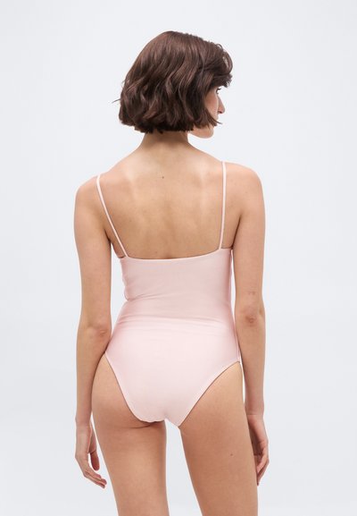 Kobieta z krótkimi brązowymi włosami, ubraną w jasnoróżowy body na cienkich ramiączkach, pokazana od tyłu na jednolitym białym tle.