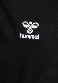 Prenda de algodón negra con un logotipo bordado en blanco de una abeja y el texto "hummel" abajo, mostrando una textura suave y un diseño sencillo.