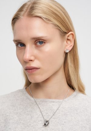 Blonde vrouw met blauwe ogen draagt een lichtgrijze top, een zilveren ketting met een gedraaide hanger en ronde zilveren studs.