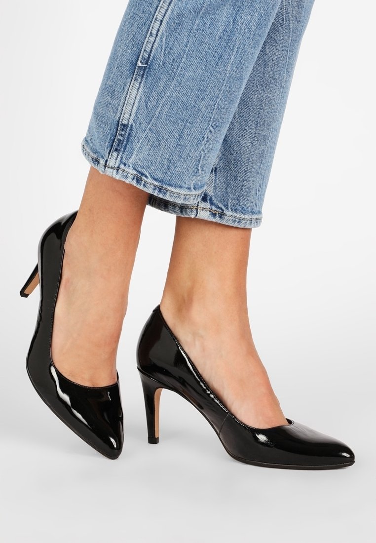 Pieds portant des escarpins à talons hauts en cuir verni noir brillant associés à un jean court en denim bleu clair sur fond blanc.