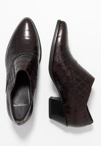 Burgundy ankelboots med krokodilsk texture, med spetsig tå och låg blockklack, placerade mot en vit bakgrund.