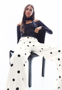 Fekete vállvillantós felső és fehér, fekete pöttyös, bő szárú nadrág. A modell egy fekete széken ül, világos háttér előtt.