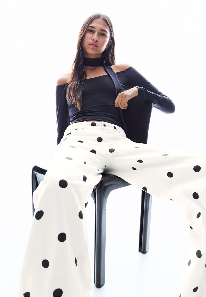Fekete vállvillantós felső és fehér, fekete pöttyös, bő szárú nadrág. A modell egy fekete széken ül, világos háttér előtt.