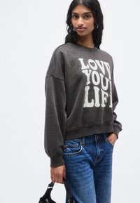 Σκούρο γκρι φούτερ με την επιγραφή «LOVE YOU LIFE» σε άσπρο, με χαλαρή εφαρμογή, ριμπ μανσέτες και τελείωμα. Συνδυάζεται με μπλε τζιν.